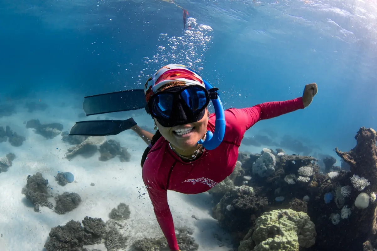 Hannah smiling underwater in a red rashie above a coral reef