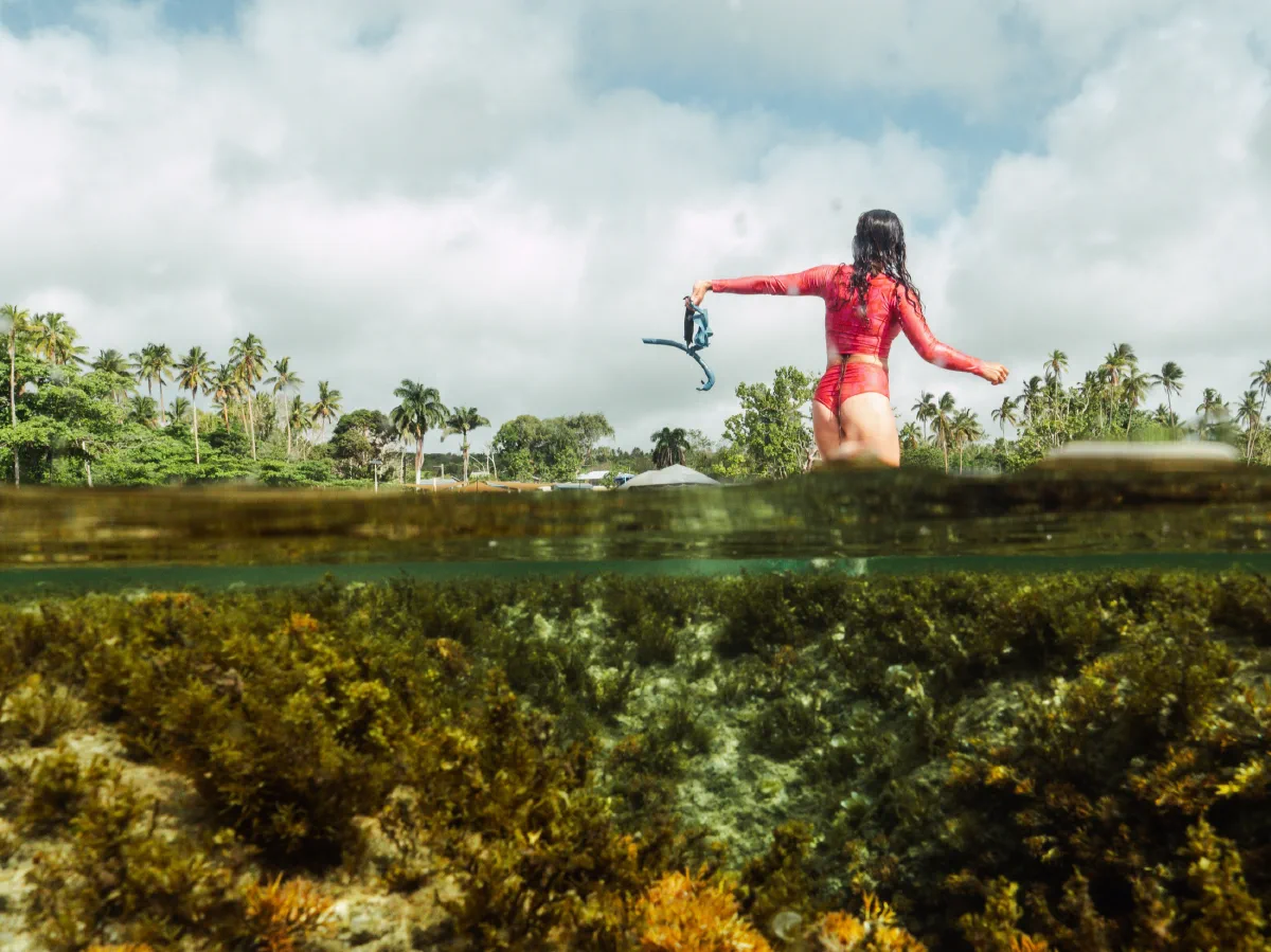 Tee holding her snorkel above the water, with reef and a palm-lined shore in a split-level view